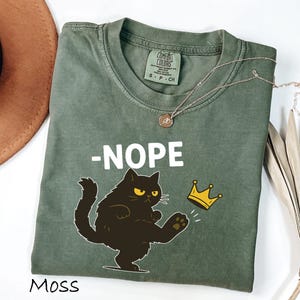 May include: Moss green t-shirt with the word "-NOPE" in white block letters and a cartoon black cat kicking a gold crown. The shirt has a Comfort Colors tag and a round neckline. A gold necklace is draped across the shirt.