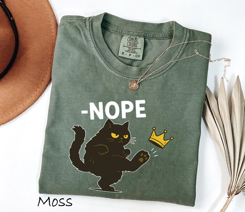 May include: Moss green t-shirt with the word "-NOPE" in white block letters and a cartoon black cat kicking a gold crown. The shirt has a Comfort Colors tag and a round neckline. A gold necklace is draped across the shirt.