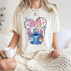 May include: Cream-coloured t-shirt featuring a graphic of the Disney character Stitch, with a castle and bow design. The shirt is a relaxed fit, with short sleeves. The design includes pink, blue, and yellow accents. The shirt is worn by a person.