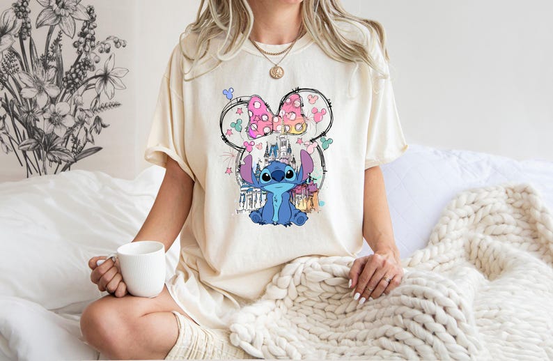 May include: Cream-coloured t-shirt featuring a graphic of the Disney character Stitch, with a castle and bow design. The shirt is a relaxed fit, with short sleeves. The design includes pink, blue, and yellow accents. The shirt is worn by a person.