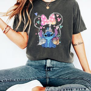 May include: Dark grey t-shirt with a graphic of Stitch from Lilo & Stitch, a pink bow, and a castle design. The design is within a Mickey Mouse ear outline. The shirt is worn with blue jeans and white trainers.