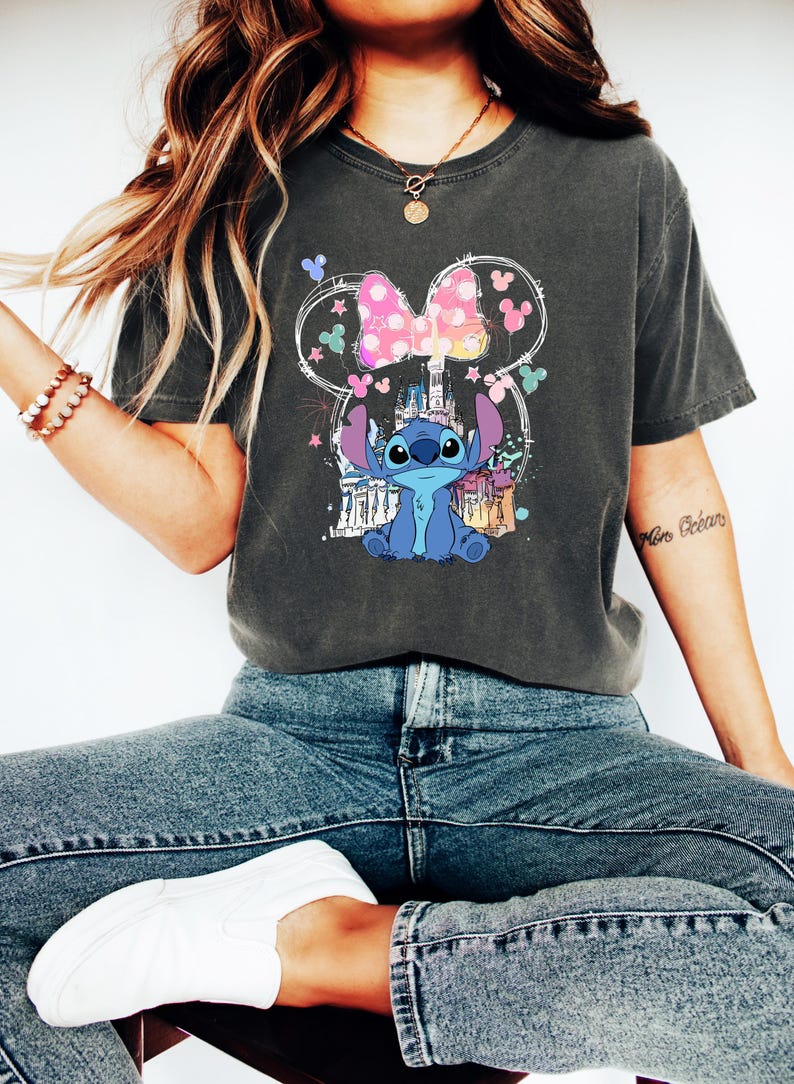 May include: Dark grey t-shirt with a graphic of Stitch from Lilo & Stitch, a pink bow, and a castle design. The design is within a Mickey Mouse ear outline. The shirt is worn with blue jeans and white trainers.