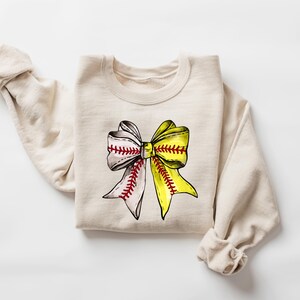 May include: A beige sweatshirt with a yellow and white softball bow graphic. The bow is tied with red stitching.