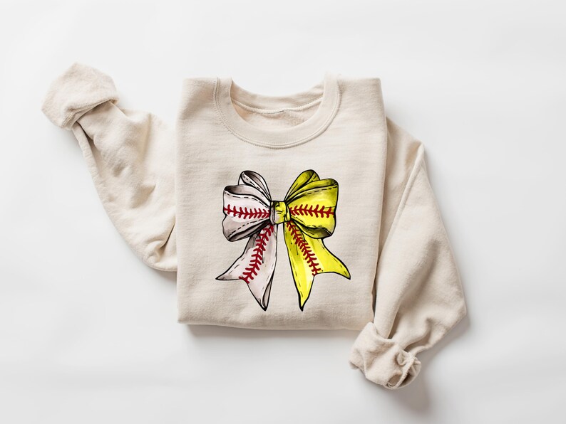May include: A beige sweatshirt with a yellow and white softball bow graphic. The bow is tied with red stitching.