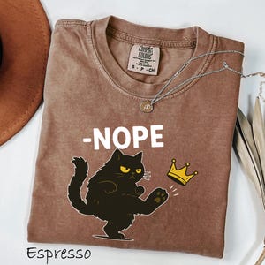 May include: Espresso-colored t-shirt with the word "-NOPE" in white block letters. A black cat with yellow eyes is kicking a gold crown. The shirt has a crew neck and a Comfort Colors tag.