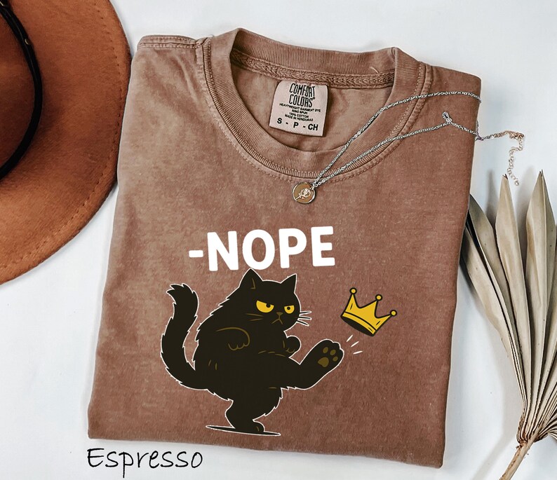 May include: Espresso-colored t-shirt with the word "-NOPE" in white block letters. A black cat with yellow eyes is kicking a gold crown. The shirt has a crew neck and a Comfort Colors tag.