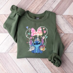 May include: An olive green sweatshirt with a graphic of Stitch from Lilo & Stitch. The design includes a Minnie Mouse-style bow, a castle, and other colourful elements. The sweatshirt is made of a soft material.