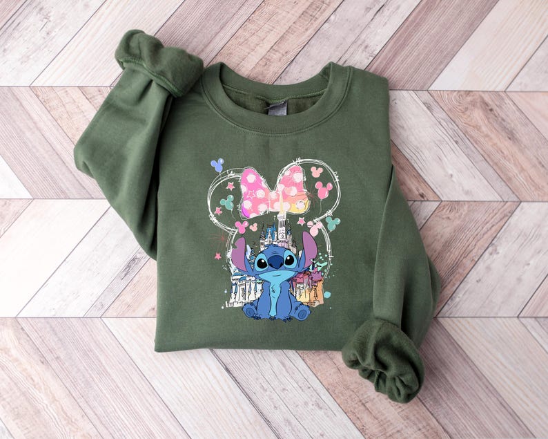 May include: An olive green sweatshirt with a graphic of Stitch from Lilo & Stitch. The design includes a Minnie Mouse-style bow, a castle, and other colourful elements. The sweatshirt is made of a soft material.