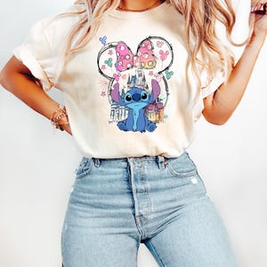 May include: Cream-coloured t-shirt featuring a graphic of Stitch from Lilo & Stitch, framed by a Minnie Mouse bow and ears. The design includes a castle and fireworks. The shirt is paired with light blue jeans.