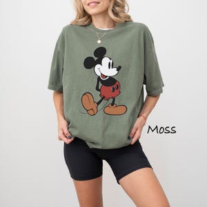 May include: Olive green t-shirt featuring a classic Mickey Mouse graphic. The cartoon character is depicted in black, white, red, and tan. The word "Moss" is printed in black on the lower right side of the image.