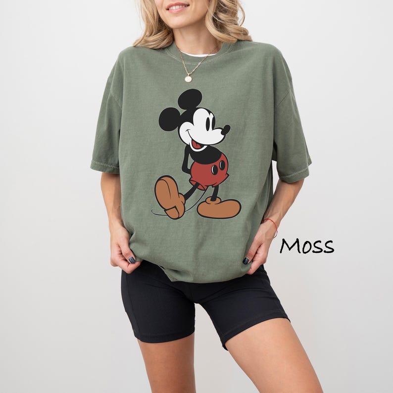 May include: Olive green t-shirt featuring a classic Mickey Mouse graphic. The cartoon character is depicted in black, white, red, and tan. The word "Moss" is printed in black on the lower right side of the image.