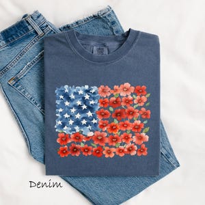 May include: A navy blue t-shirt with a watercolour design of the American flag. The flag design features white stars on a blue background and red flowers. The word "Denim" is printed at the bottom.