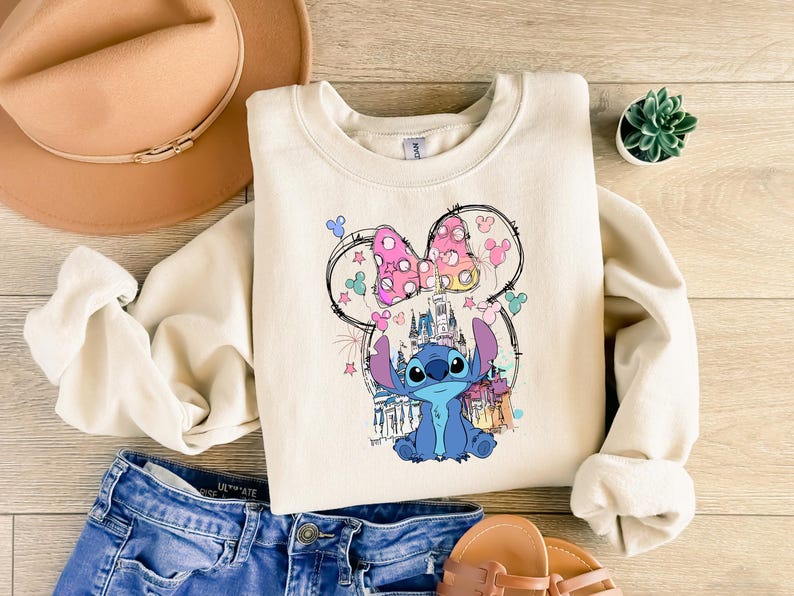 May include: A cream-coloured sweatshirt with a colourful graphic of Stitch from Lilo & Stitch, with a Minnie Mouse bow and a castle. The sweatshirt is paired with blue denim shorts, a tan hat, and sandals.