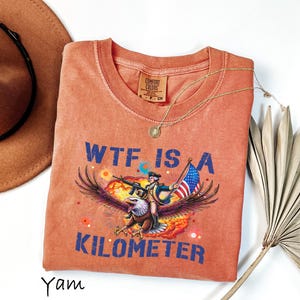 K&ouml;nnte beinhalten: Orangefarbenes Comfort Colors T-Shirt mit einem Aufdruck eines Adlers, der eine Figur mit einer Waffe und einer amerikanischen Flagge tr&auml;gt. Der Text "WTF IS A KILOMETER" ist blau gedruckt. Eine goldene Halskette und ein brauner Hut sind ebenfalls sichtbar.