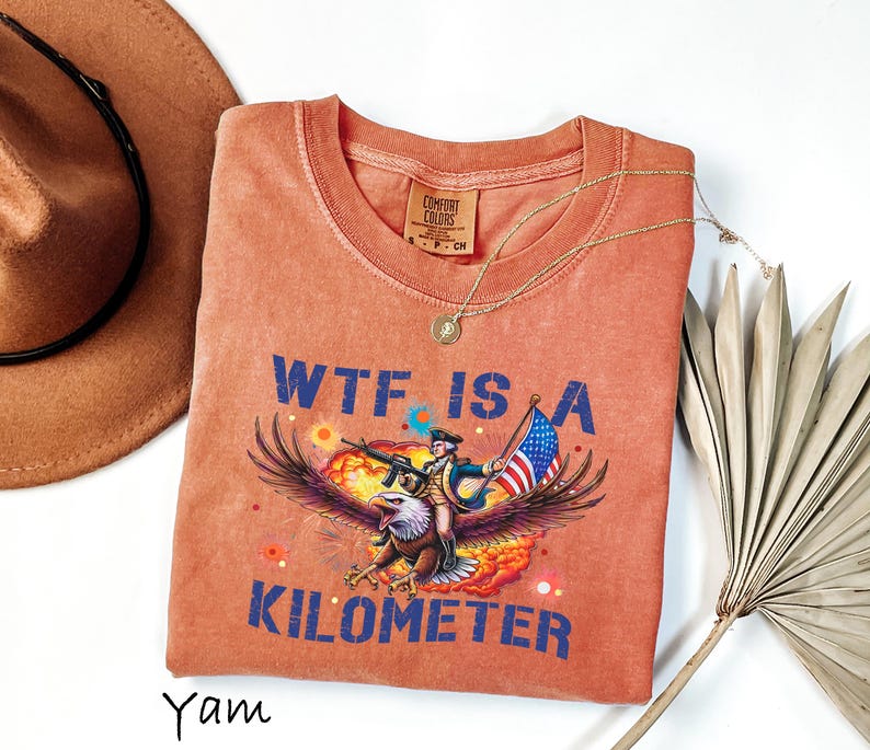 K&ouml;nnte beinhalten: Orangefarbenes Comfort Colors T-Shirt mit einem Aufdruck eines Adlers, der eine Figur mit einer Waffe und einer amerikanischen Flagge tr&auml;gt. Der Text "WTF IS A KILOMETER" ist blau gedruckt. Eine goldene Halskette und ein brauner Hut sind ebenfalls sichtbar.