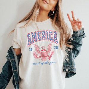 May include: White t-shirt with a blue and red graphic of an eagle with the words "AMERICA 17 76 land of the free" printed on it.
