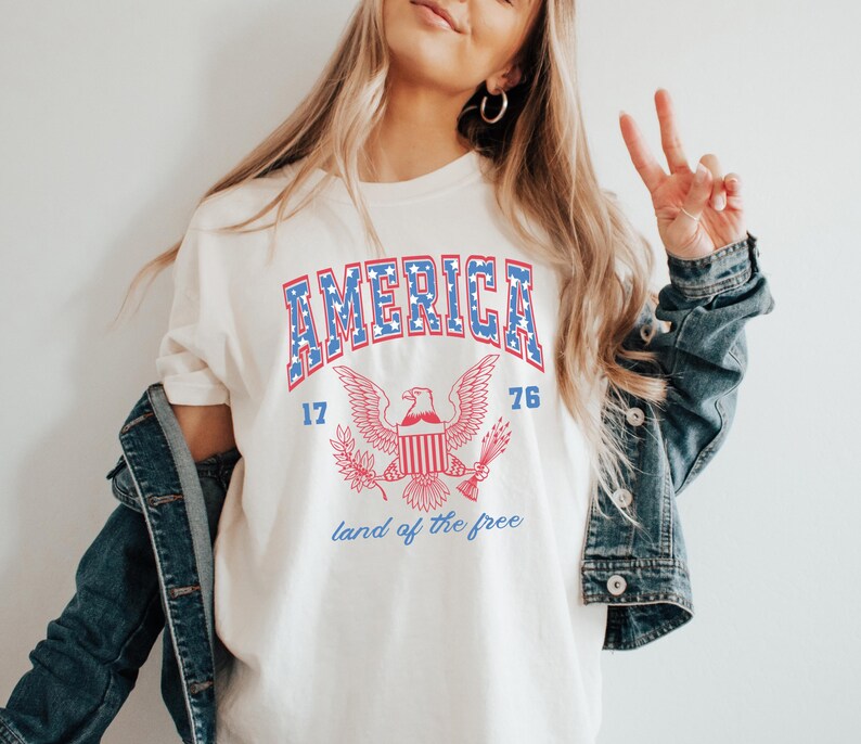 May include: White t-shirt with a blue and red graphic of an eagle with the words "AMERICA 17 76 land of the free" printed on it.