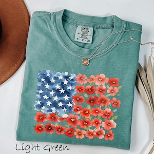 May include: Light green t-shirt with a floral American flag design. The flag features blue watercolor stars and red flowers. The shirt has a crew neck and short sleeves. The text "Light Green" is at the bottom of the image.
