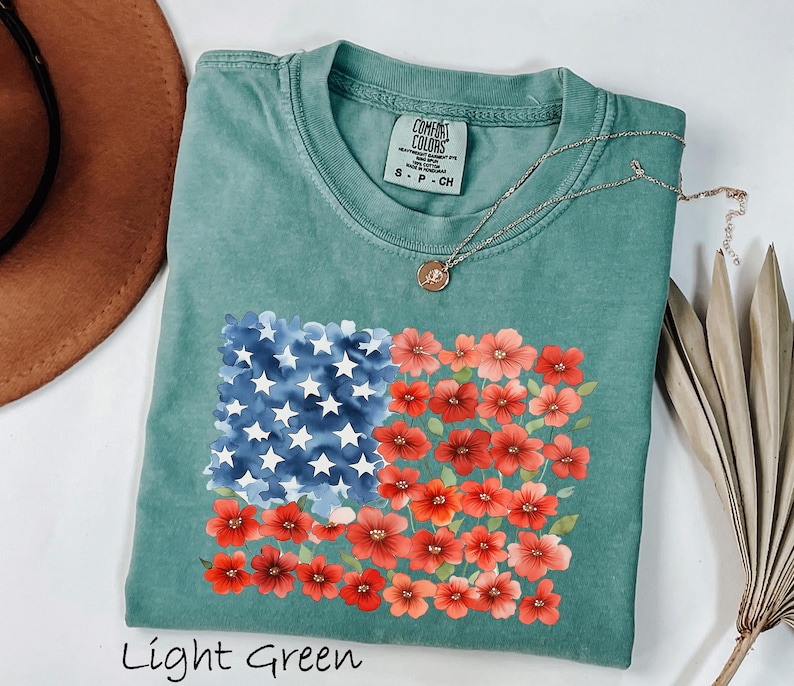 May include: Light green t-shirt with a floral American flag design. The flag features blue watercolor stars and red flowers. The shirt has a crew neck and short sleeves. The text "Light Green" is at the bottom of the image.