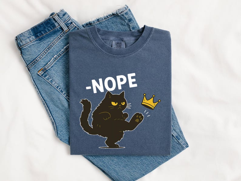 May include: A navy blue t-shirt with the word "-NOPE" in white, and a cartoon black cat kicking a gold crown. The shirt is laid on top of a pair of blue jeans.