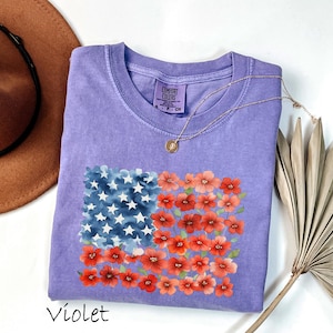 May include: Violet-coloured t-shirt with a floral American flag design. The flag design incorporates blue watercolour stars and red watercolour flowers. A gold necklace with a pendant is draped on the shirt. A brown hat and dried palm leaf are in the background.