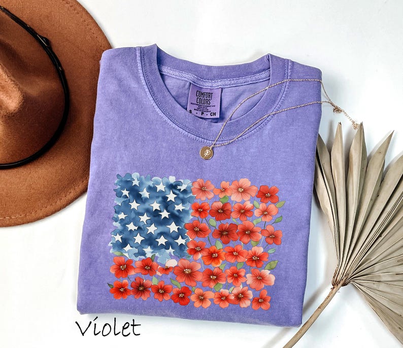May include: Violet-coloured t-shirt with a floral American flag design. The flag design incorporates blue watercolour stars and red watercolour flowers. A gold necklace with a pendant is draped on the shirt. A brown hat and dried palm leaf are in the background.