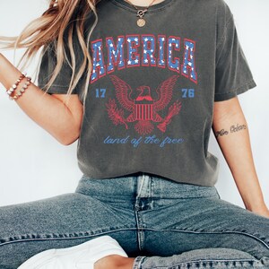 May include: A gray t-shirt with a vintage-style graphic of an eagle and the word "AMERICA" in blue with white stars. The eagle is holding a banner that says "land of the free". The numbers "17" and "76" are on either side of the eagle.