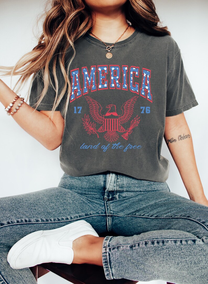 May include: A gray t-shirt with a vintage-style graphic of an eagle and the word "AMERICA" in blue with white stars. The eagle is holding a banner that says "land of the free". The numbers "17" and "76" are on either side of the eagle.