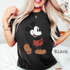 May include: Black t-shirt featuring a classic Mickey Mouse graphic. The cartoon character is depicted in his signature colors: black, red, white, and brown. The word "Black" is printed in white on the lower right side of the shirt.