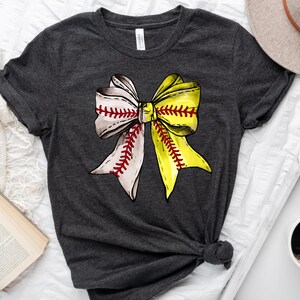May include: A dark gray t-shirt with a softball-themed bow design. The bow is made of white and yellow fabric with red stitching, resembling a softball.