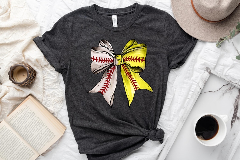 May include: A dark gray t-shirt with a softball-themed bow design. The bow is made of white and yellow fabric with red stitching, resembling a softball.