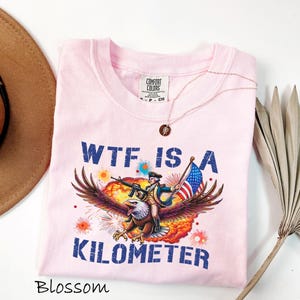 K&ouml;nnte beinhalten: Hellrosa T-Shirt mit einer Grafik eines Adlers, der eine Person mit einer Waffe und einer amerikanischen Flagge tr&auml;gt. Der Text "WTF IS A KILOMETER" ist blau gedruckt. Eine goldene Halskette befindet sich auf dem T-Shirt. Das T-Shirt ist mit "Blossom" beschriftet.
