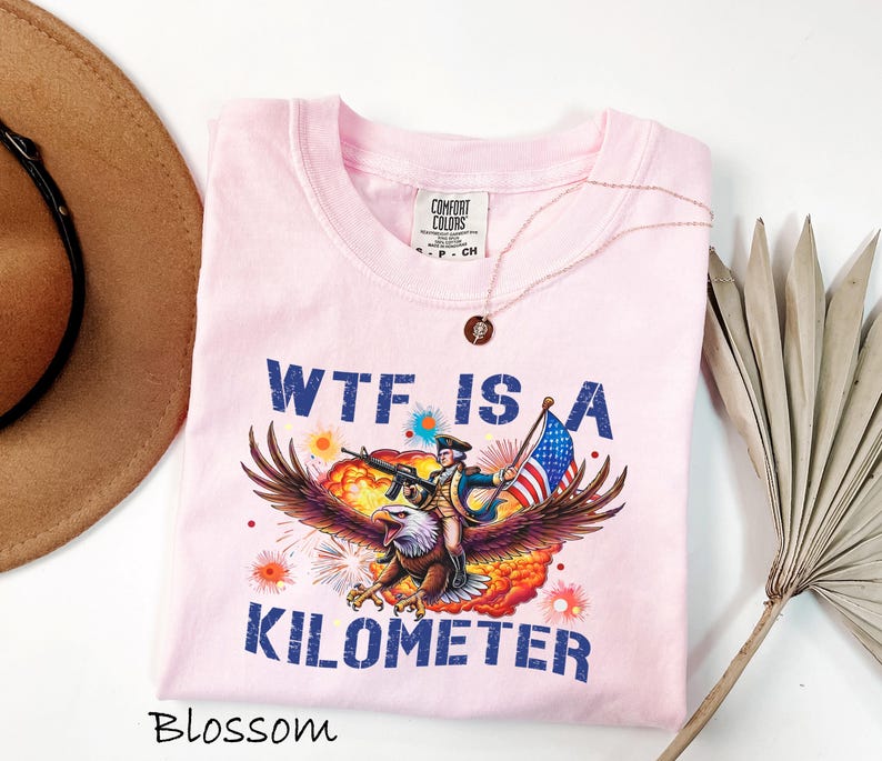 K&ouml;nnte beinhalten: Hellrosa T-Shirt mit einer Grafik eines Adlers, der eine Person mit einer Waffe und einer amerikanischen Flagge tr&auml;gt. Der Text "WTF IS A KILOMETER" ist blau gedruckt. Eine goldene Halskette befindet sich auf dem T-Shirt. Das T-Shirt ist mit "Blossom" beschriftet.