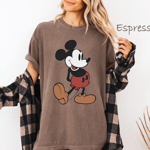 May include: Espresso-colored t-shirt featuring a classic Mickey Mouse graphic. The cartoon character is depicted in black, white, and red. The shirt has rolled-up sleeves and is worn with a black and brown plaid shirt.