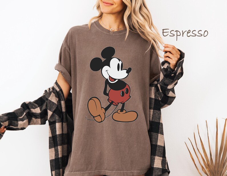 May include: Espresso-colored t-shirt featuring a classic Mickey Mouse graphic. The cartoon character is depicted in black, white, and red. The shirt has rolled-up sleeves and is worn with a black and brown plaid shirt.