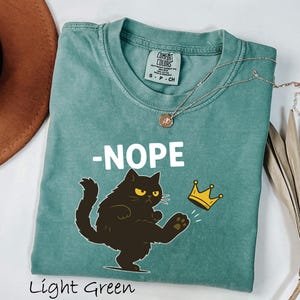 May include: Light green t-shirt with a graphic of a black cat kicking a gold crown, with the word "-NOPE" in white. The shirt is made of a soft material and has a round neckline. A silver necklace with a pendant is on the shirt.
