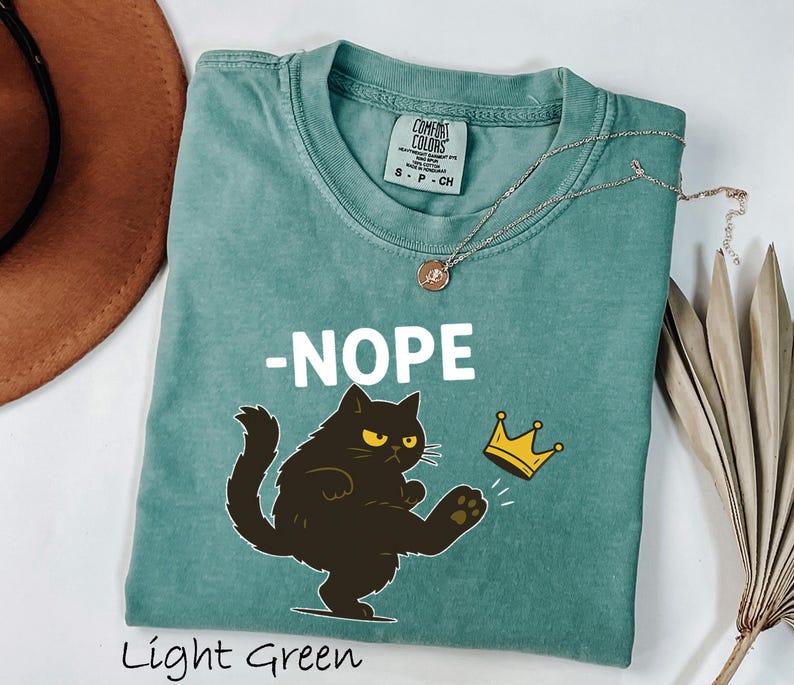 May include: Light green t-shirt with a graphic of a black cat kicking a gold crown, with the word "-NOPE" in white. The shirt is made of a soft material and has a round neckline. A silver necklace with a pendant is on the shirt.