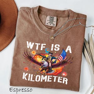 K&ouml;nnte beinhalten: Espressofarbenes T-Shirt mit einer Grafik eines Adlers, der einen Soldaten mit Gewehr und amerikanischer Flagge tr&auml;gt. Der Text "WTF IS A KILOMETER" ist wei&szlig; gedruckt. Eine silberne Halskette liegt &uuml;ber dem Shirt.