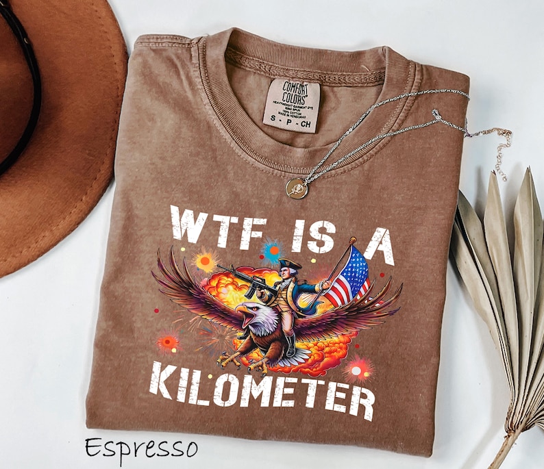 K&ouml;nnte beinhalten: Espressofarbenes T-Shirt mit einer Grafik eines Adlers, der einen Soldaten mit Gewehr und amerikanischer Flagge tr&auml;gt. Der Text "WTF IS A KILOMETER" ist wei&szlig; gedruckt. Eine silberne Halskette liegt &uuml;ber dem Shirt.