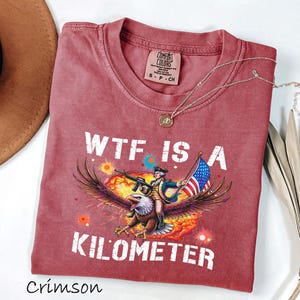K&ouml;nnte beinhalten: Karmesinrotes T-Shirt mit dem Text "WTF IS A KILOMETER" und einer Grafik eines Adlers, der einen Soldaten mit Gewehr und amerikanischer Flagge tr&auml;gt. Das Shirt hat einen Rundhalsausschnitt und kurze &Auml;rmel. Eine goldene Halskette liegt &uuml;ber dem Shirt.