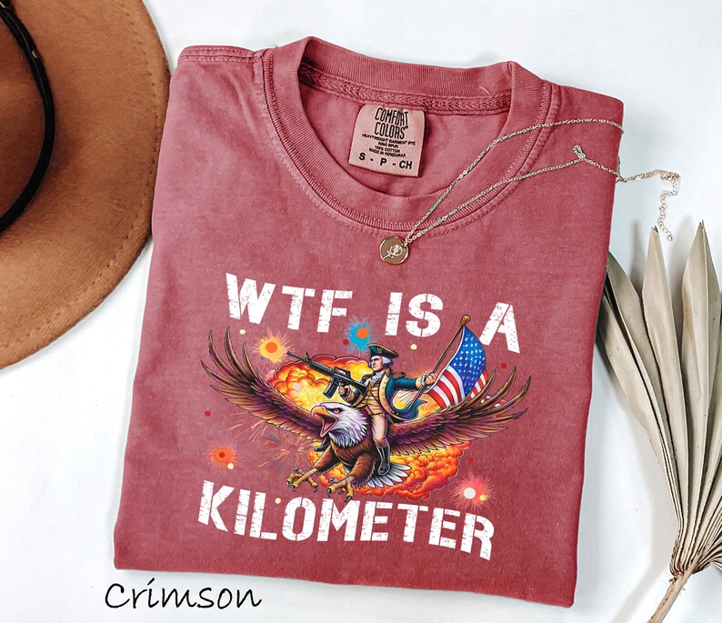 K&ouml;nnte beinhalten: Karmesinrotes T-Shirt mit dem Text "WTF IS A KILOMETER" und einer Grafik eines Adlers, der einen Soldaten mit Gewehr und amerikanischer Flagge tr&auml;gt. Das Shirt hat einen Rundhalsausschnitt und kurze &Auml;rmel. Eine goldene Halskette liegt &uuml;ber dem Shirt.