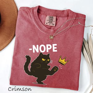 May include: Crimson-colored t-shirt with a graphic of a black cat kicking a gold crown, with the word "-NOPE" in white. A gold necklace with a pendant is on the shirt. The shirt is labeled "Comfort Colors".