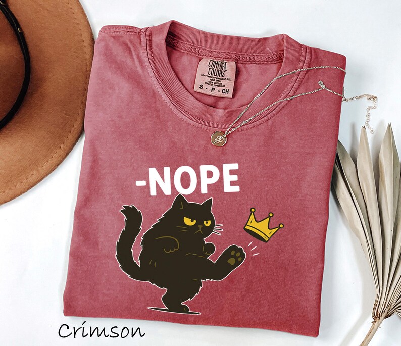 May include: Crimson-colored t-shirt with a graphic of a black cat kicking a gold crown, with the word "-NOPE" in white. A gold necklace with a pendant is on the shirt. The shirt is labeled "Comfort Colors".