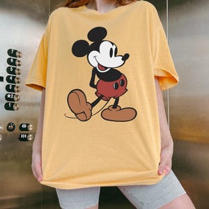 May include: Yellow t-shirt featuring a classic Mickey Mouse graphic. The cartoon character is depicted in black, white, red, and brown. The shirt has short sleeves and a relaxed fit, perfect for casual wear.