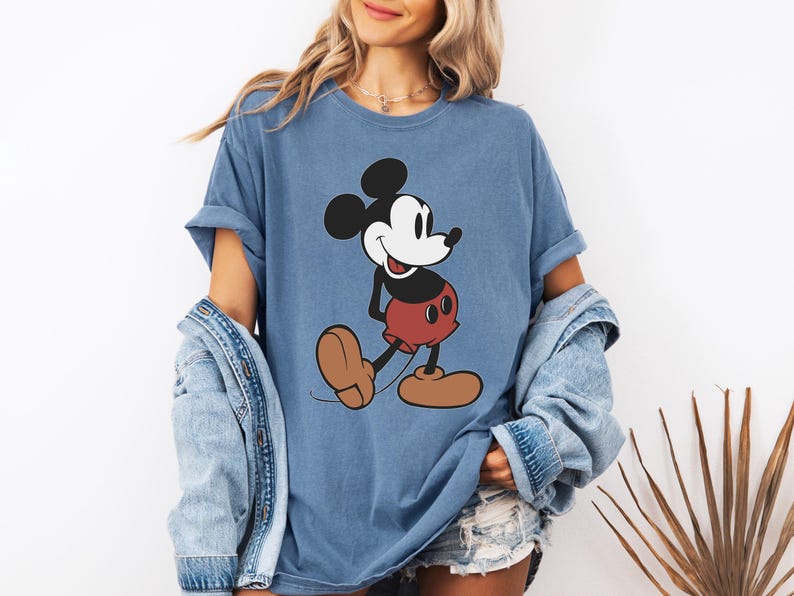 May include: A blue t-shirt featuring a classic cartoon character, Mickey Mouse. The graphic shows Mickey in his iconic pose, with black ears, a white face, red shorts, and brown shoes. The shirt has rolled-up sleeves.