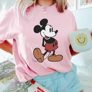 May include: A light pink t-shirt featuring a large graphic of Mickey Mouse in his classic pose. The cartoon character is depicted in black, white, brown, and red. The person is also holding a white mug with a yellow smiley face.