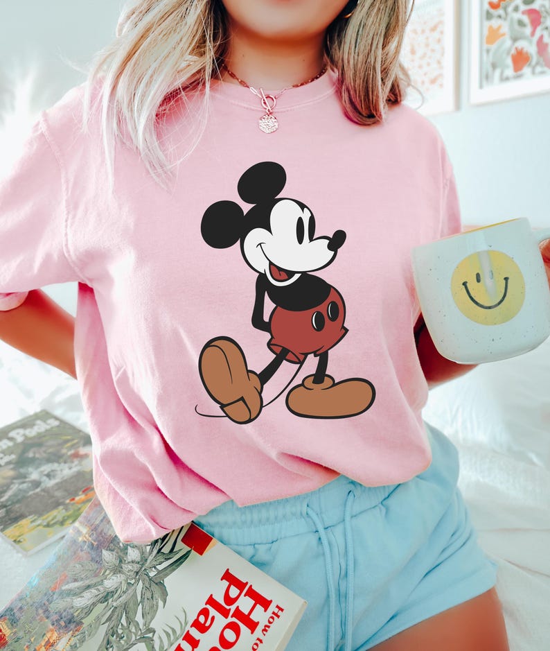 May include: A light pink t-shirt featuring a large graphic of Mickey Mouse in his classic pose. The cartoon character is depicted in black, white, brown, and red. The person is also holding a white mug with a yellow smiley face.