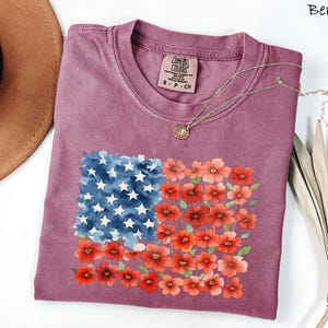 May include: Berry-coloured t-shirt with a floral American flag design. The flag design features blue watercolour stars and red and orange flowers. The shirt is a Comfort Colors brand, size S-P-CH.