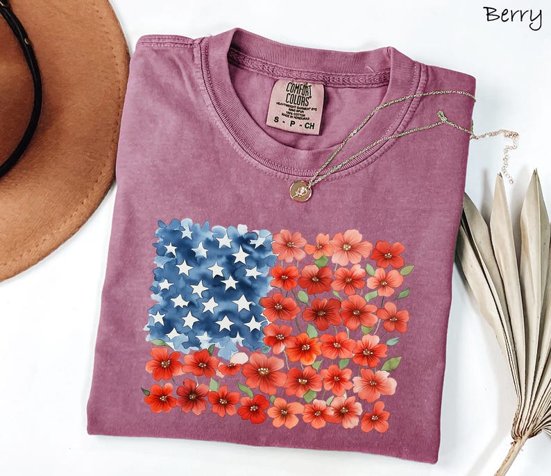 May include: Berry-coloured t-shirt with a floral American flag design. The flag design features blue watercolour stars and red and orange flowers. The shirt is a Comfort Colors brand, size S-P-CH.