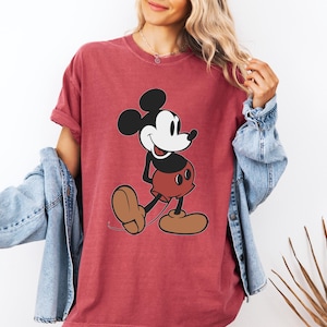 May include: Red t-shirt featuring a classic cartoon Mickey Mouse design. The graphic shows Mickey in his iconic pose, with black ears, a white face, red shorts, and brown shoes. The shirt is a solid red color.